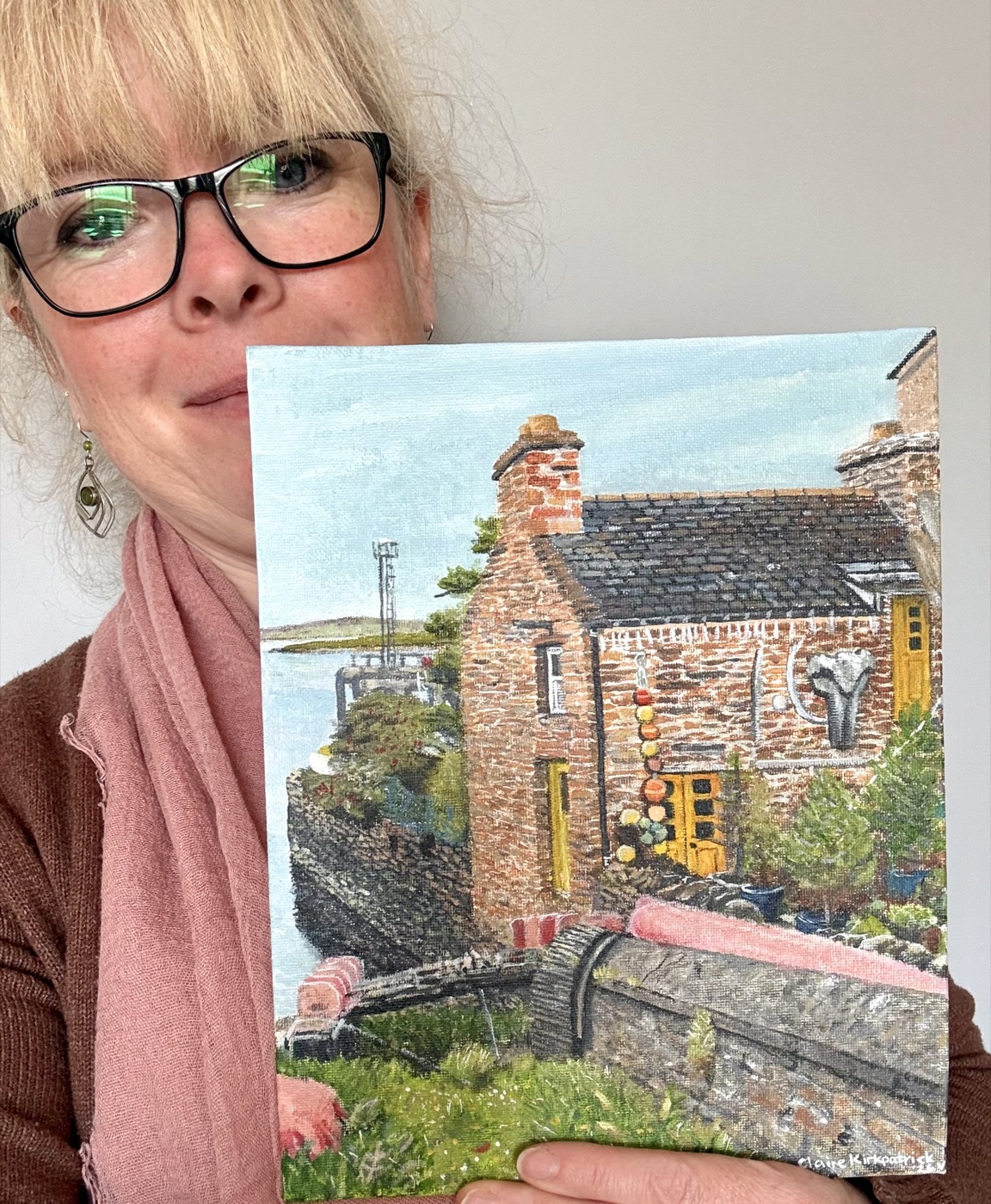 Claire Kirkpatrick holding one of her paintings of an Orkney cottage by the water